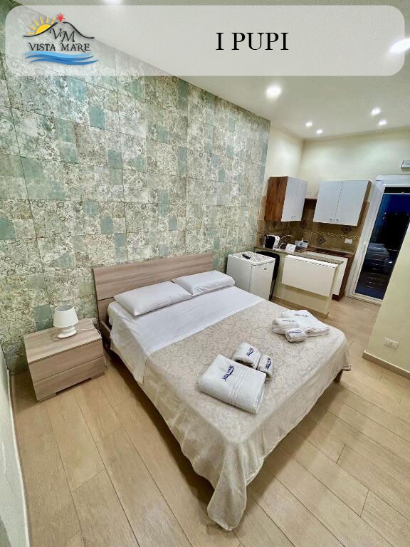 A cozy bedroom is presented with a spacious bed, featuring light bedding and neatly arranged towels. The walls are decorated with intricate green tiles, complementing the light wood furniture. A small kitchenette and dining area are visible in the background.