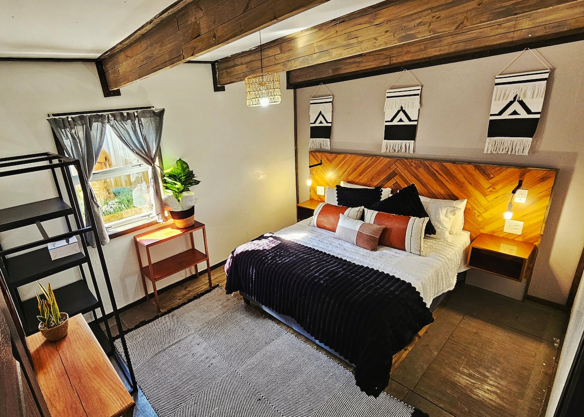 A well-appointed bedroom features a spacious bed with dark bedding and a wooden headboard. Accents of orange and cream are included in the decorative pillows. Natural light enters through a window dressed with sheer curtains, while a green plant adds a touch of greenery on a bedside table.