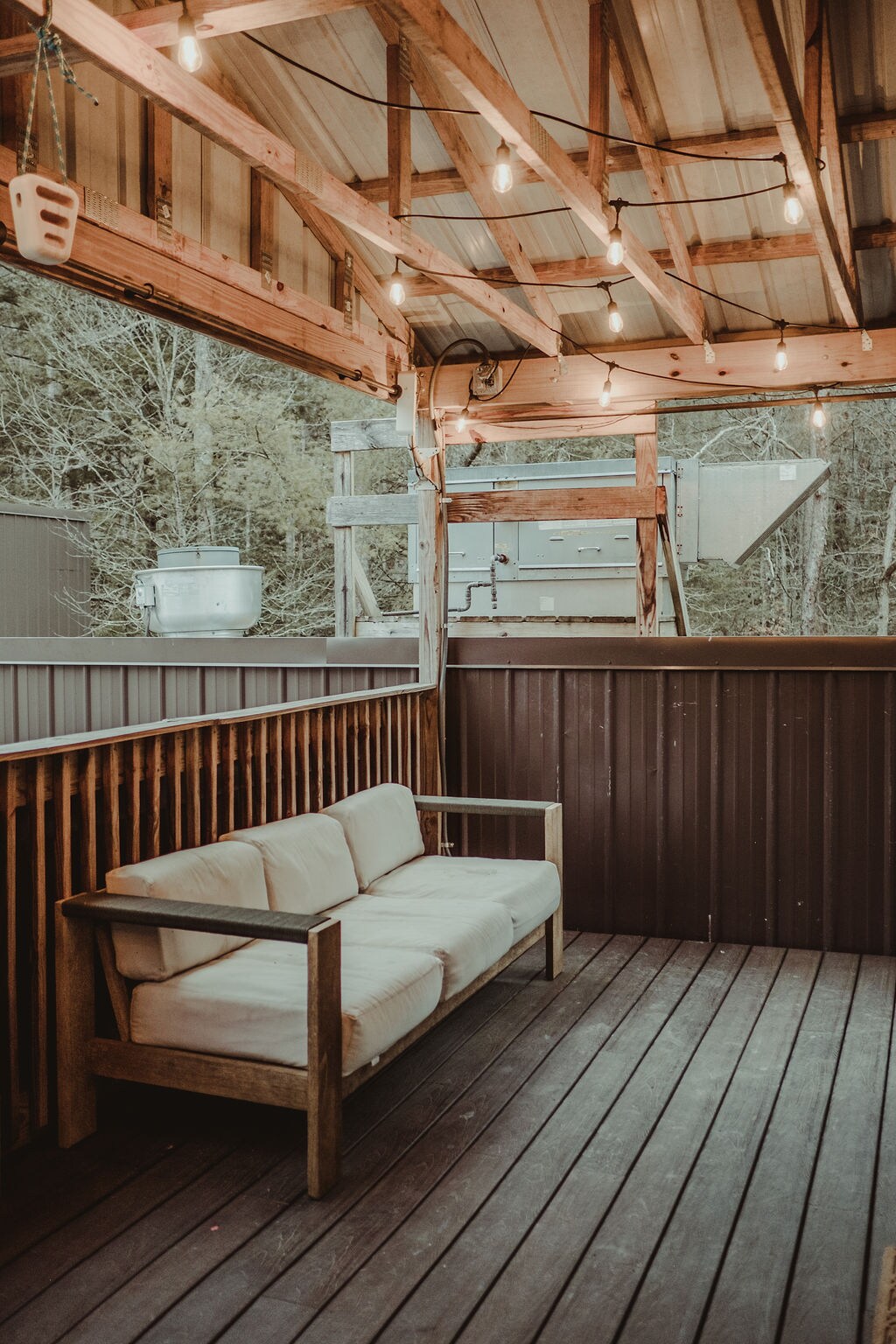 An outdoor seating area features a wooden bench with light, comfortable cushions. String lights hang overhead, providing soft illumination. The deck showcases wooden flooring and a partially enclosed space, surrounded by natural greenery in the backdrop.
