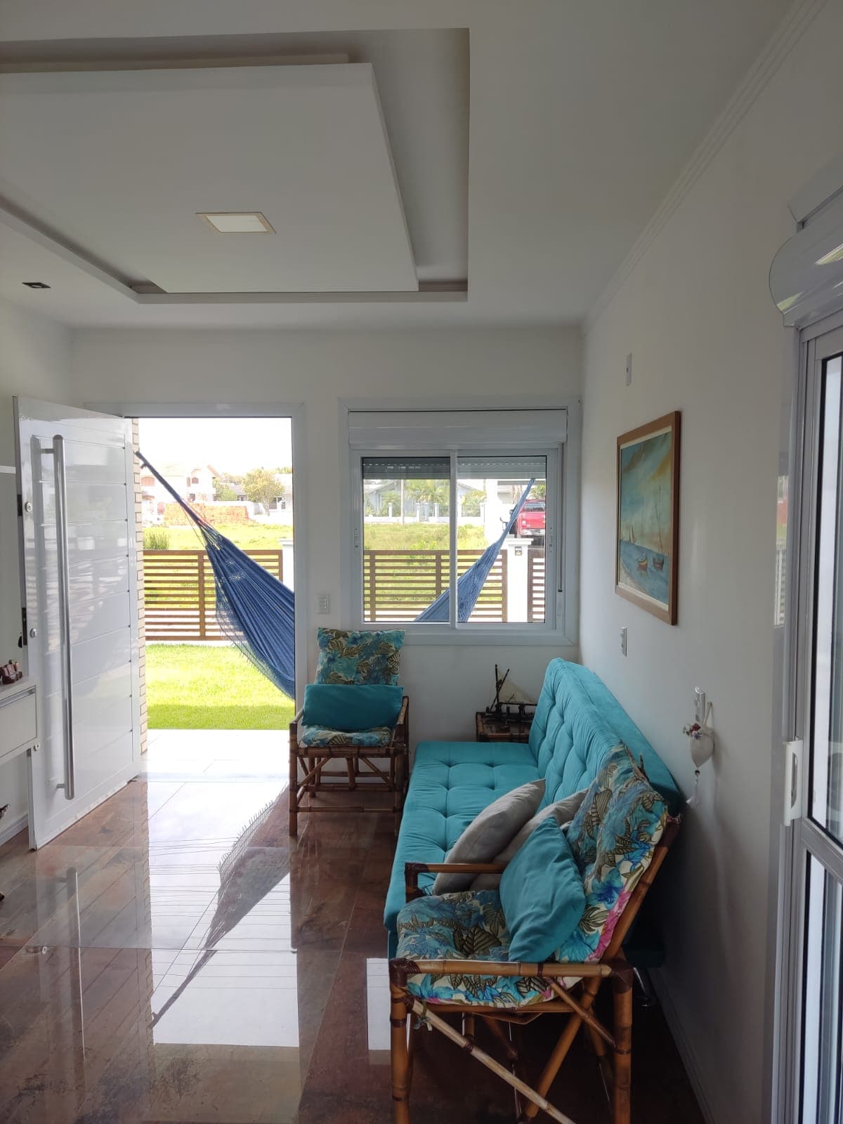 A welcoming living area features a comfortable turquoise sofa and two chairs with floral patterns. Natural light pours in through large windows, illuminating the space. A hammock is visible, creating an inviting spot for relaxation. The flooring is polished, enhancing the room's brightness.
