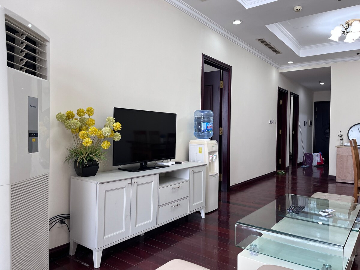 A comfortably arranged living room features a white television stand with a flat-screen TV and a decorative flower arrangement. A water cooler and a glass coffee table are visible, while a glimpse of a hallway with wooden flooring and doorways can be seen in the background.