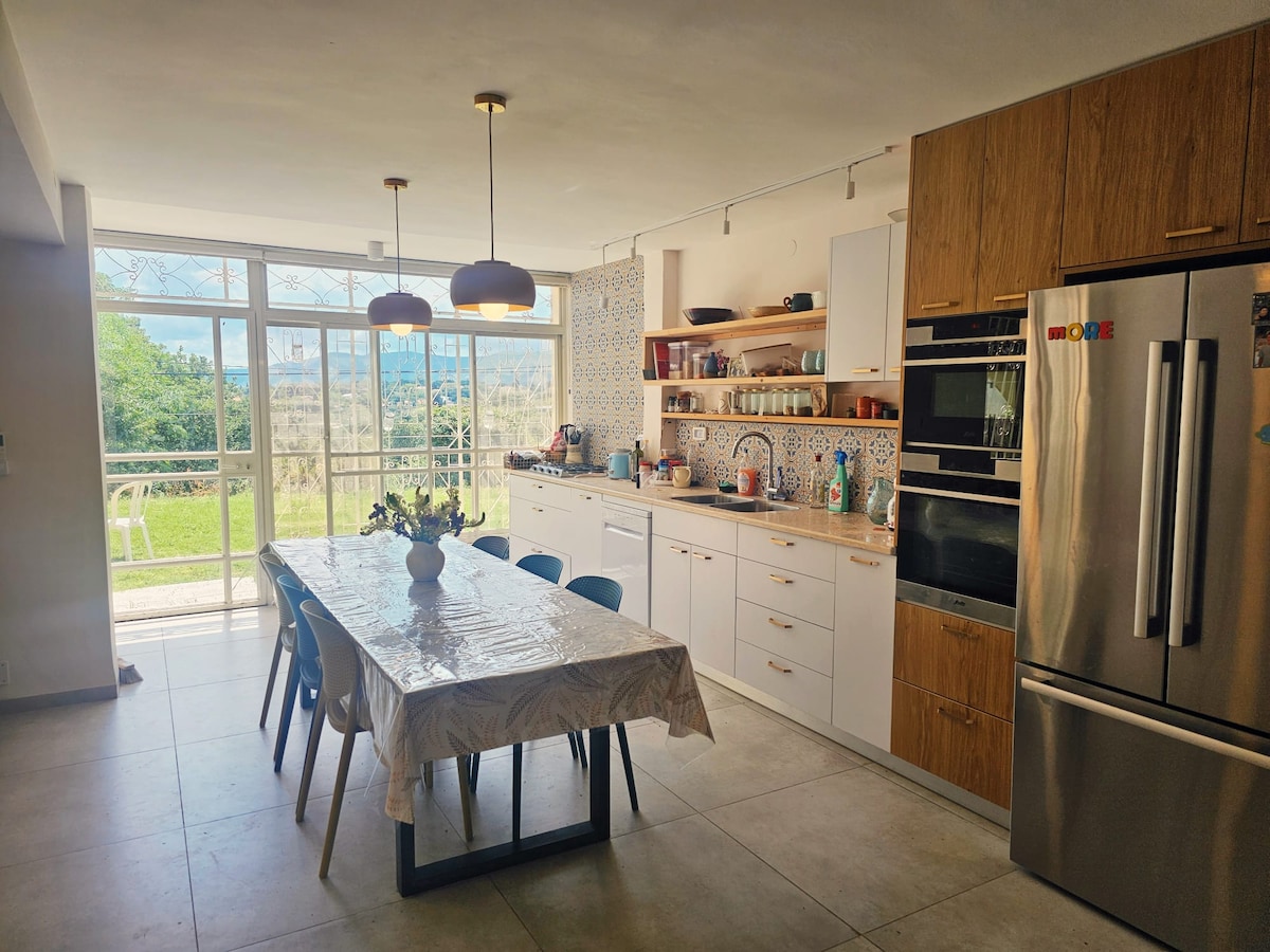 The kitchen and dining area features a spacious table surrounded by six chairs, with natural light streaming in through large glass doors. The countertop is equipped with modern appliances, and decorative tiles can be seen along the backsplash. A view of the greenery outside enhances the inviting atmosphere.