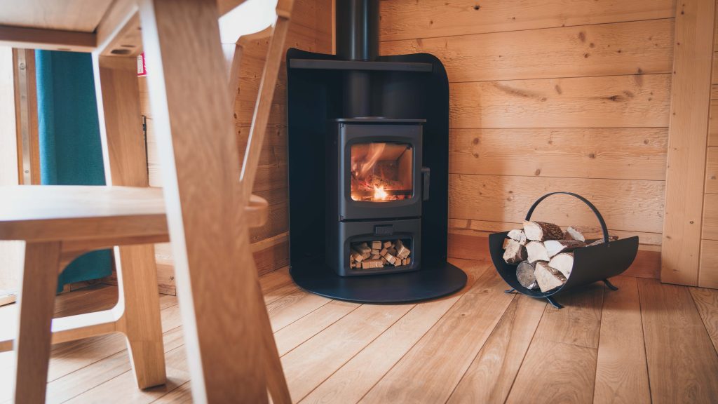 A wood-burning stove is positioned in a cozy corner, surrounded by warm wooden walls. A stack of logs rests in a metal holder, enhancing the rustic ambiance. The natural wooden floor complements the inviting atmosphere of the space.