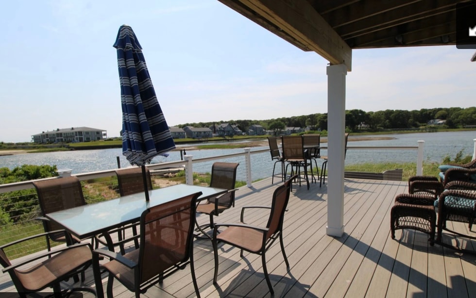 An outdoor dining area is presented on a spacious deck, featuring a large table surrounded by several metal chairs. A blue and white striped umbrella provides shade, while a serene water view and lush green surroundings add to the serene ambiance.