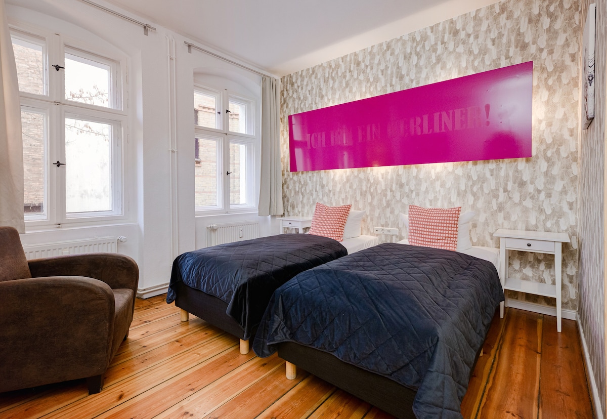 The room features two separate beds with navy-colored bedding and decorative pillows. Light hardwood flooring complements wallpaper with a subtle pattern. A large, bright panel with text adds a unique design element. Natural light enters through two windows, providing a welcoming atmosphere.