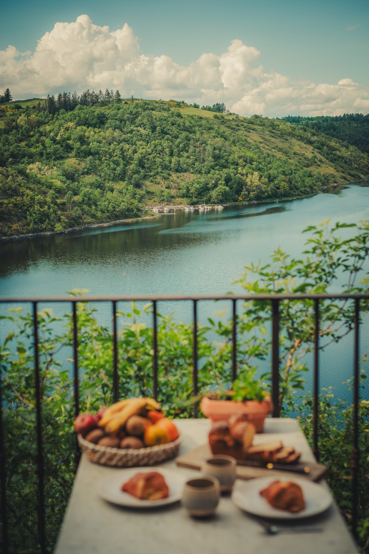 A scenic view of the lake and lush green hills is presented from a balcony. A table is set with plates of fresh pastries and fruits, accompanied by cups, inviting a calm and relaxing atmosphere.
