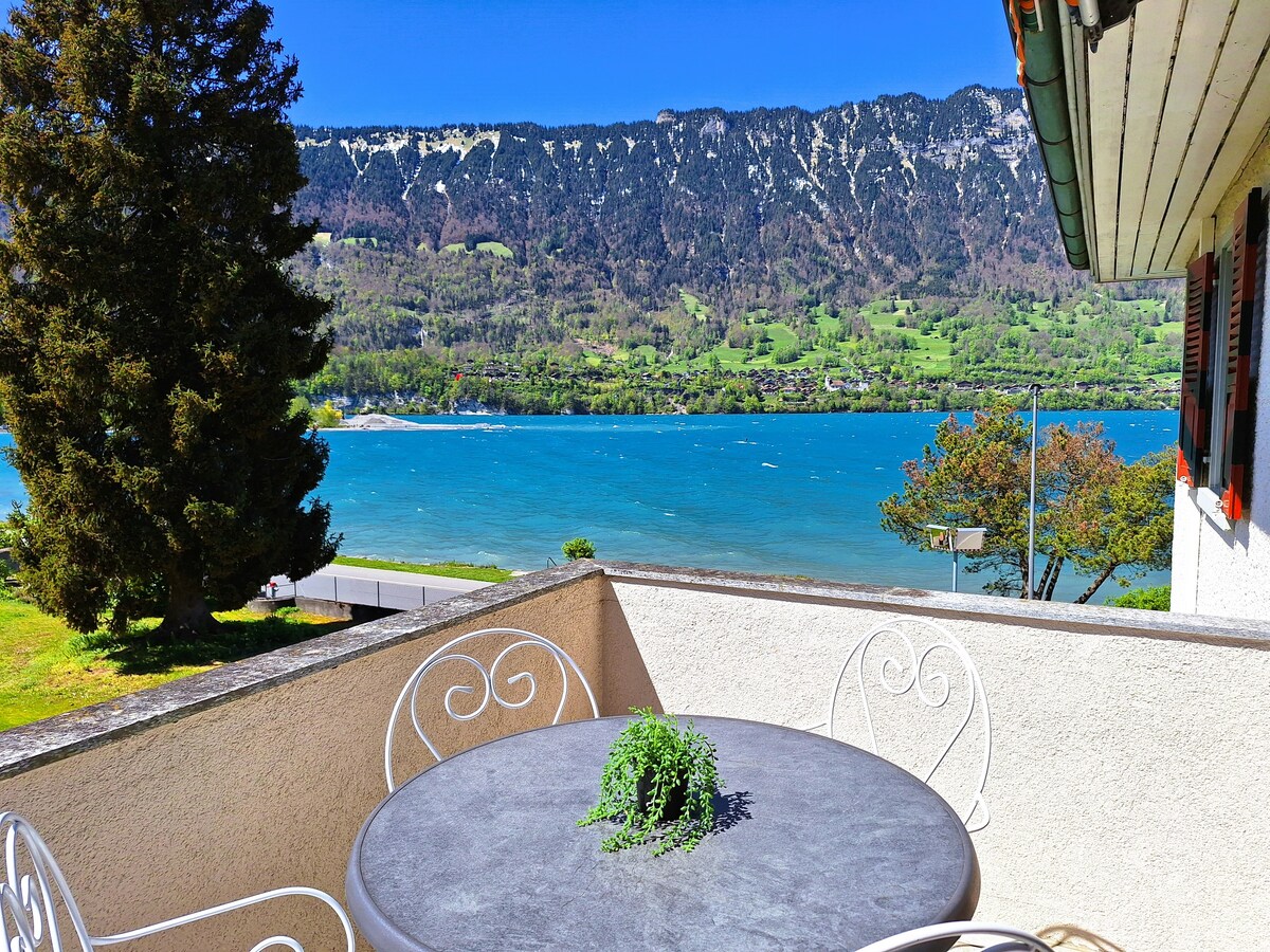 A balcony space is presented, featuring a round stone table adorned with a small potted plant. Scenic views of Lake Brienz and a mountainous landscape are visible, showcasing clear blue waters and lush greenery in the background.
