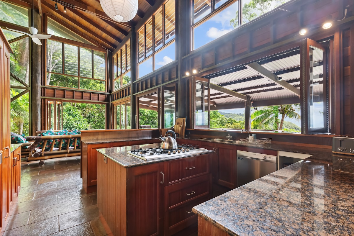 The kitchen is designed with wood cabinetry and granite countertops, showcasing a gas stove and stainless-steel appliances. Large windows provide views of the lush surroundings, and an open layout integrates dining and living areas. Natural light fills the space, enhancing the connection with nature.