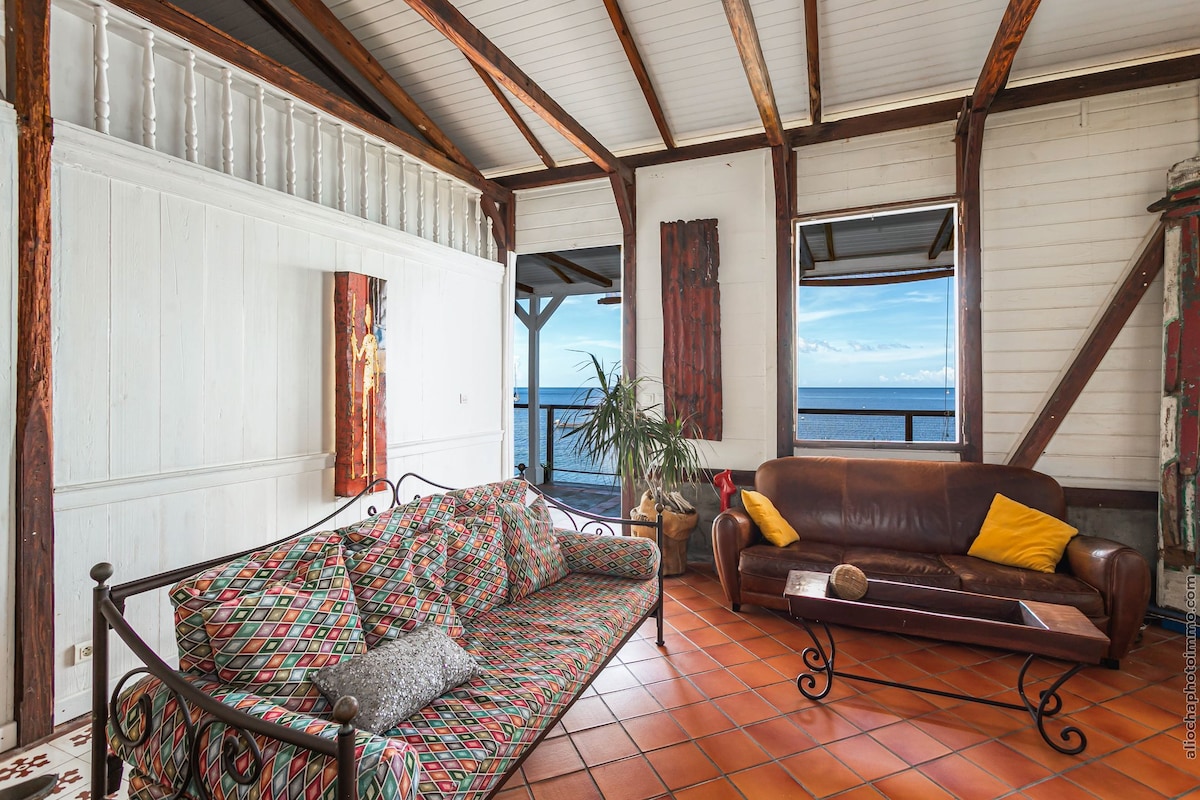 A bright living area features two distinct seating options, including a patterned sofa and a dark leather couch with a yellow throw pillow. Large windows frame views of the sea, while wooden beams and terracotta tiles add character to the space.