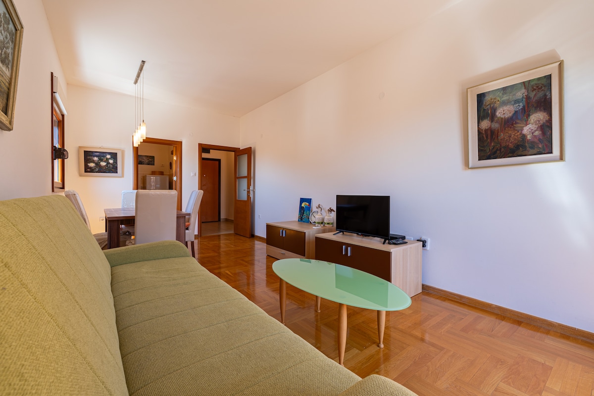 A spacious living area is shown, featuring a green sofa opposite a modern flat-screen television. A glass coffee table sits in front of the sofa, while a dining area is visible in the background with light-colored chairs. Natural light streams in through nearby windows, enhancing the inviting ambiance.