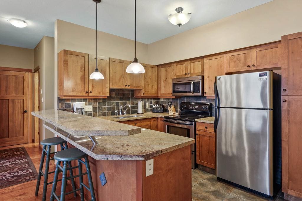 The kitchen features warm wooden cabinetry and a spacious countertop that includes three bar stools. Modern appliances, such as a stainless-steel refrigerator and oven, are visible, complemented by tile backsplash that adds texture to the area.