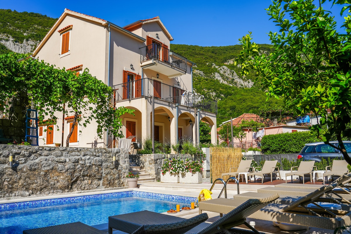 A two-story villa is depicted, surrounded by lush greenery and natural stone walls. A blue swimming pool is visible in the foreground, with sun loungers positioned nearby. The building features balconies with wooden railings, and the landscape is complemented by a mountainous backdrop.