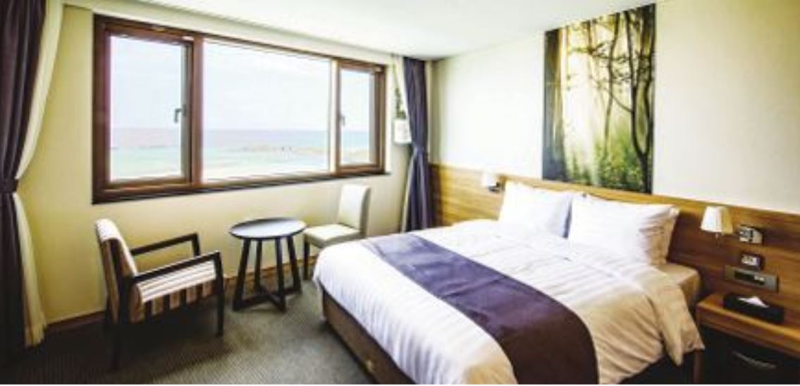 A bright guest room features a spacious bed with crisp white linens. A large window offers a view of the ocean, while a small table and two chairs add functionality. Natural light enhances the serene atmosphere, complemented by calming wall art.