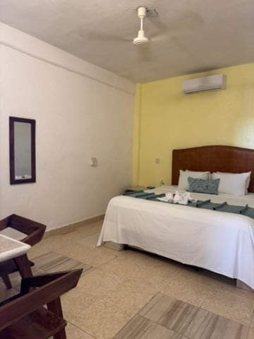 A comfortable room features a spacious bed with white linens and a decorative pillow. A small wooden table and two chairs are positioned nearby. Yellow walls complement the room, which is equipped with a ceiling fan and air conditioning unit, providing a cozy environment.