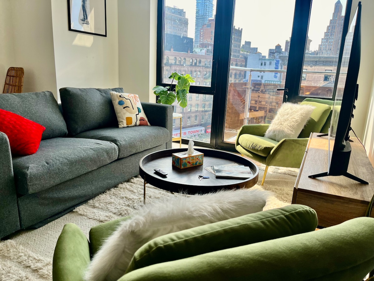 A modern living area features a gray sofa and two green armchairs, positioned near expansive windows that showcase city views. A round coffee table sits in the center, accompanied by a television and a console. Natural light enhances the cozy atmosphere.