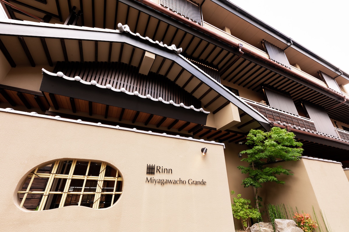 The exterior of the building features traditional architectural elements, including a sloped roof with ornamental tiles. A small tree is positioned beside the entrance, which is accentuated by large windows and a uniquely shaped cutout in the wall, offering a glimpse of the interior.