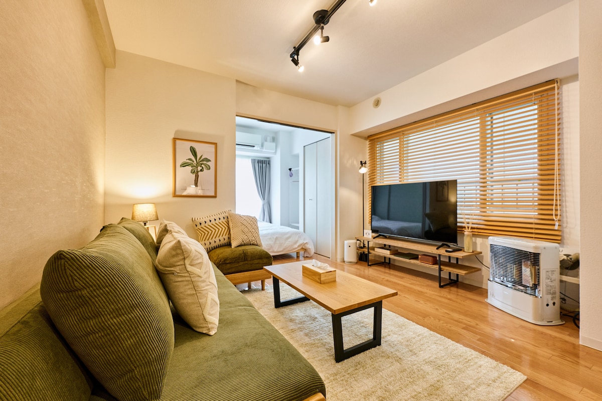 A spacious living area is illuminated by natural light from the large windows, featuring a comfortable green sofa adorned with pillows. A wooden coffee table is centered in front of a sleek television, with a warm rug underfoot enhancing the inviting layout.