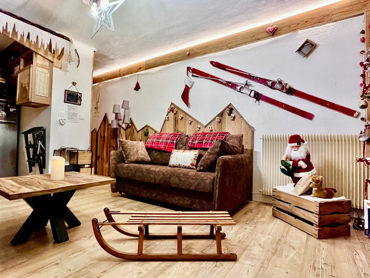 The living area features a cozy sofa adorned with festive pillows. Rustic wooden decorations line the walls, complemented by vintage skis and holiday accents. A wooden coffee table sits on the floor near a classic wooden sled, enhancing the warm atmosphere. A soft glow from a candle adds to the inviting feel.