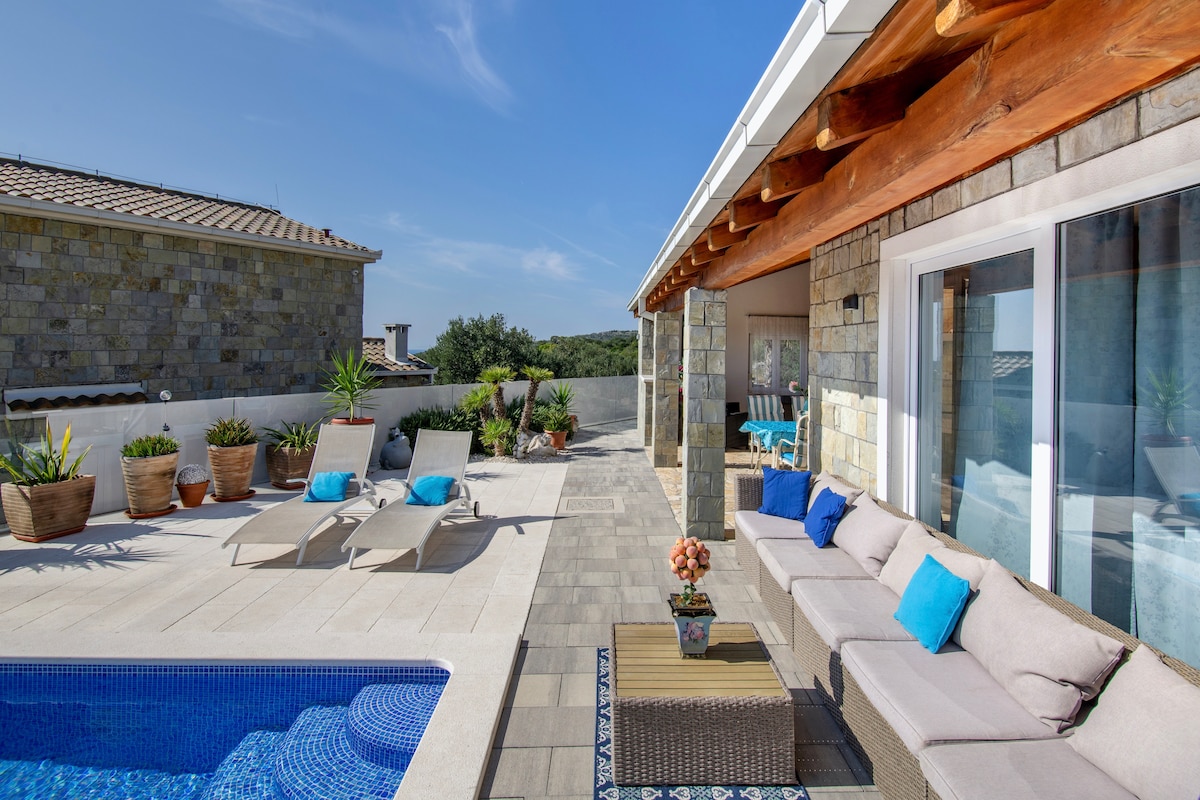 An outdoor terrace showcases a heated pool surrounded by sun loungers and potted plants. A comfortable seating area with plush cushions faces a wooden table, inviting relaxation under the clear blue sky. The villa's open design allows for unobstructed views of the scenic surroundings.
