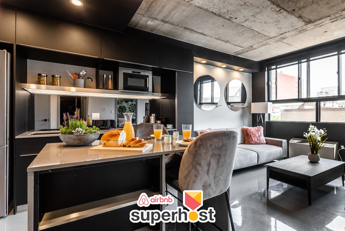 A modern kitchen and living area are featured, showcasing sleek, dark cabinetry and stainless steel appliances. A breakfast setting is visible on the countertop, with pastries and beverages. Natural light fills the space through large windows, illuminating the cozy seating arrangement.