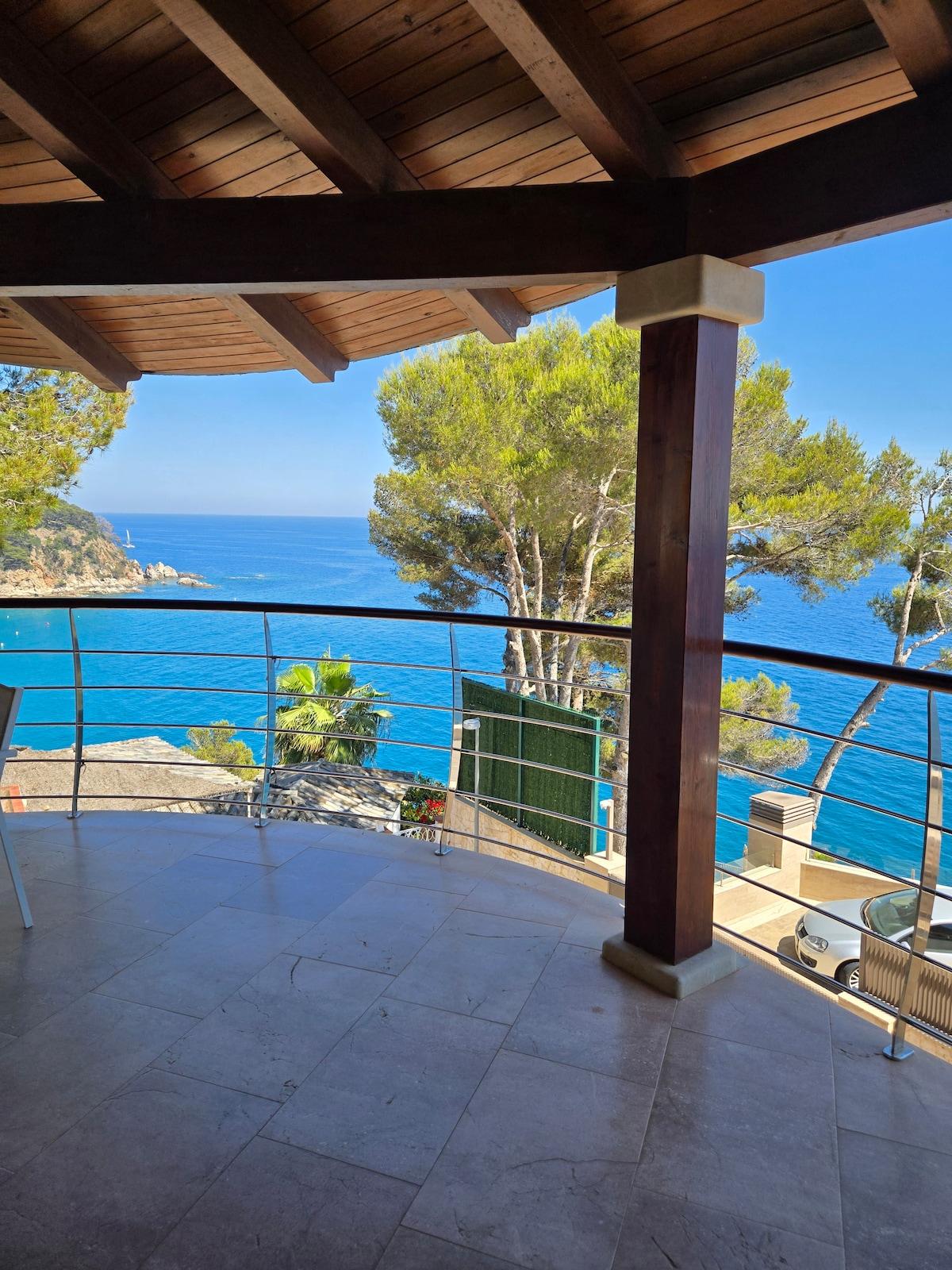 A covered terrace presents stunning views of the azure sea and surrounding greenery. A railing frames the outdoor space, which features a mix of comfortable seating options. Natural light fills the area, enhancing the spacious feeling of the open atmosphere.