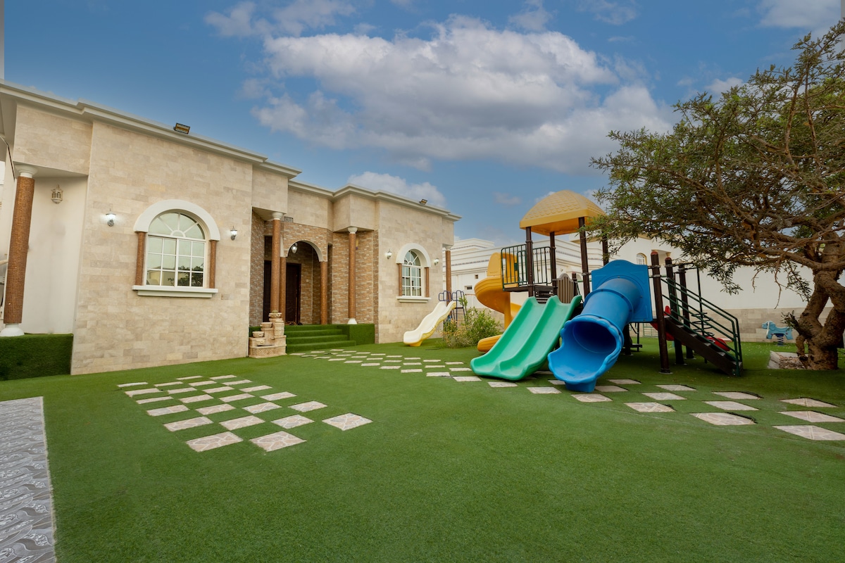 An outdoor area features a lawn with green grass, accented by a children's play structure that includes slides and climbing areas. The exterior of the house is adorned with stone and brick, complemented by arched windows under a clear blue sky.