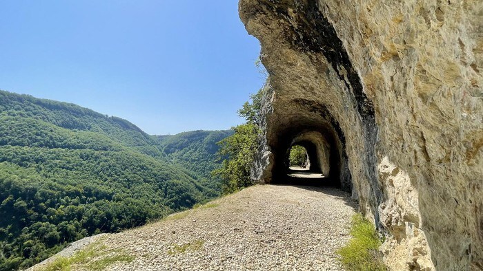 A rocky pathway is carved through a hillside, with natural arches formed by surrounding stone. Lush green trees cover the hillsides in the background, creating a serene backdrop under a clear blue sky.