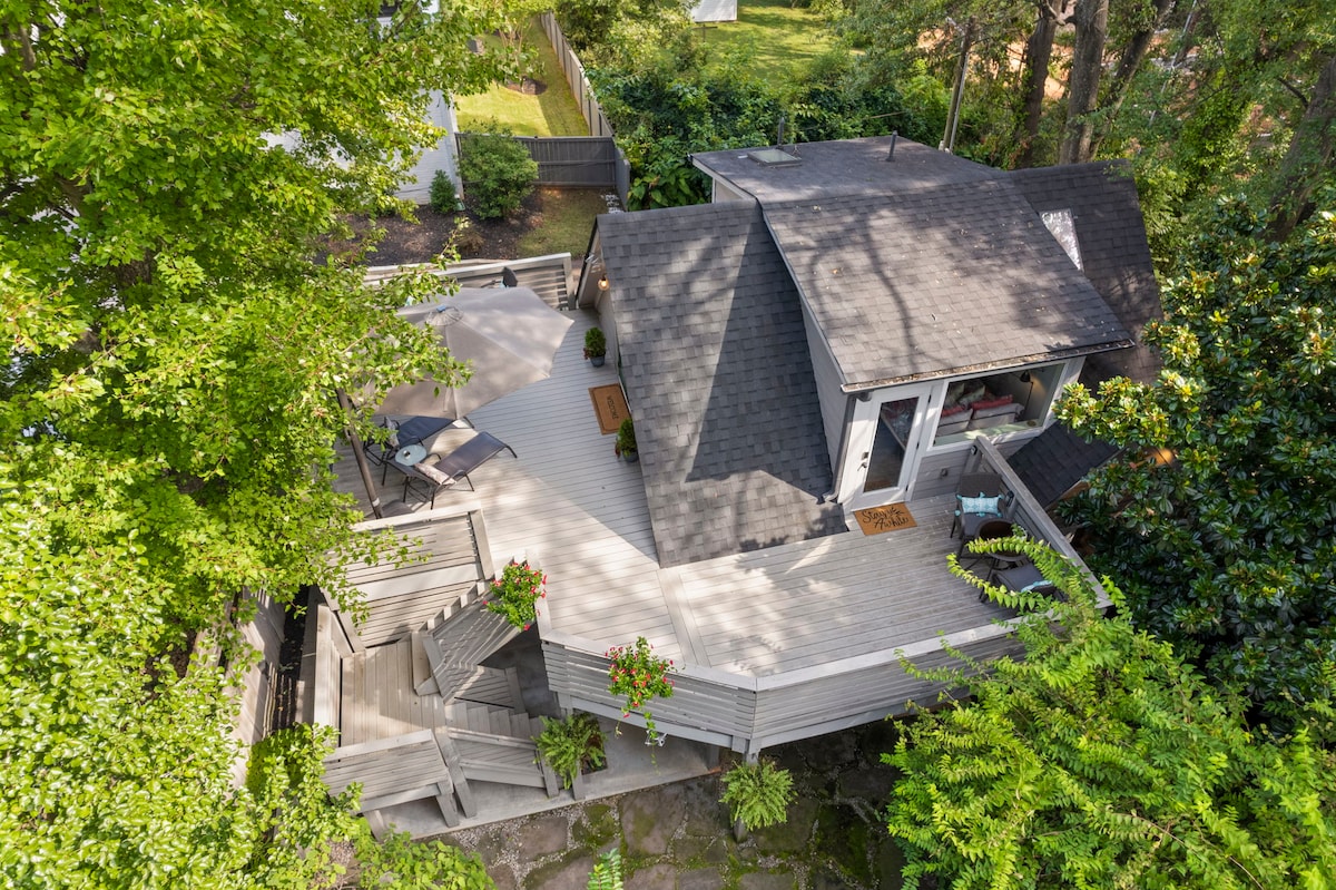 An aerial view captures the studio nestled among trees, featuring a large, multi-level deck surrounded by greenery. Comfortable seating areas are visible, and the inviting entrance showcases a mix of high-end furnishings and local decor.
