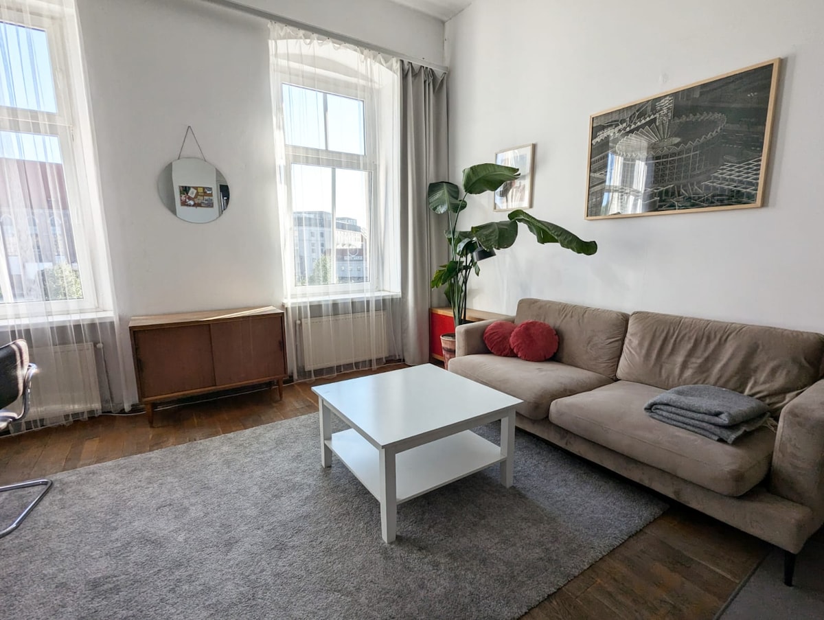 A light-filled living area features large windows draped with sheer curtains, allowing natural light to illuminate the space. A comfortable beige sofa is paired with a white coffee table, while greenery from a plant adds a touch of nature. A piece of wall art is displayed above the sofa.