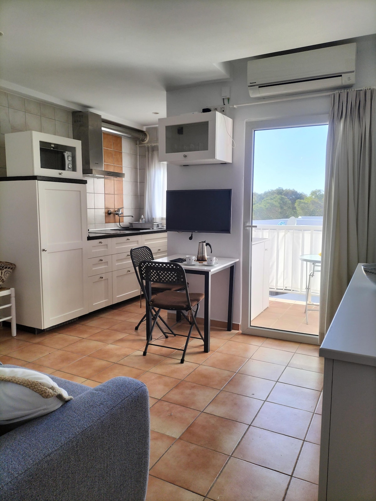 A modern kitchen space is visible, featuring white cabinetry and a compact dining area with a folding chair. A television is mounted on the wall, and a door opens to a spacious outdoor view. The room receives natural light through the window, enhancing its airy feel.