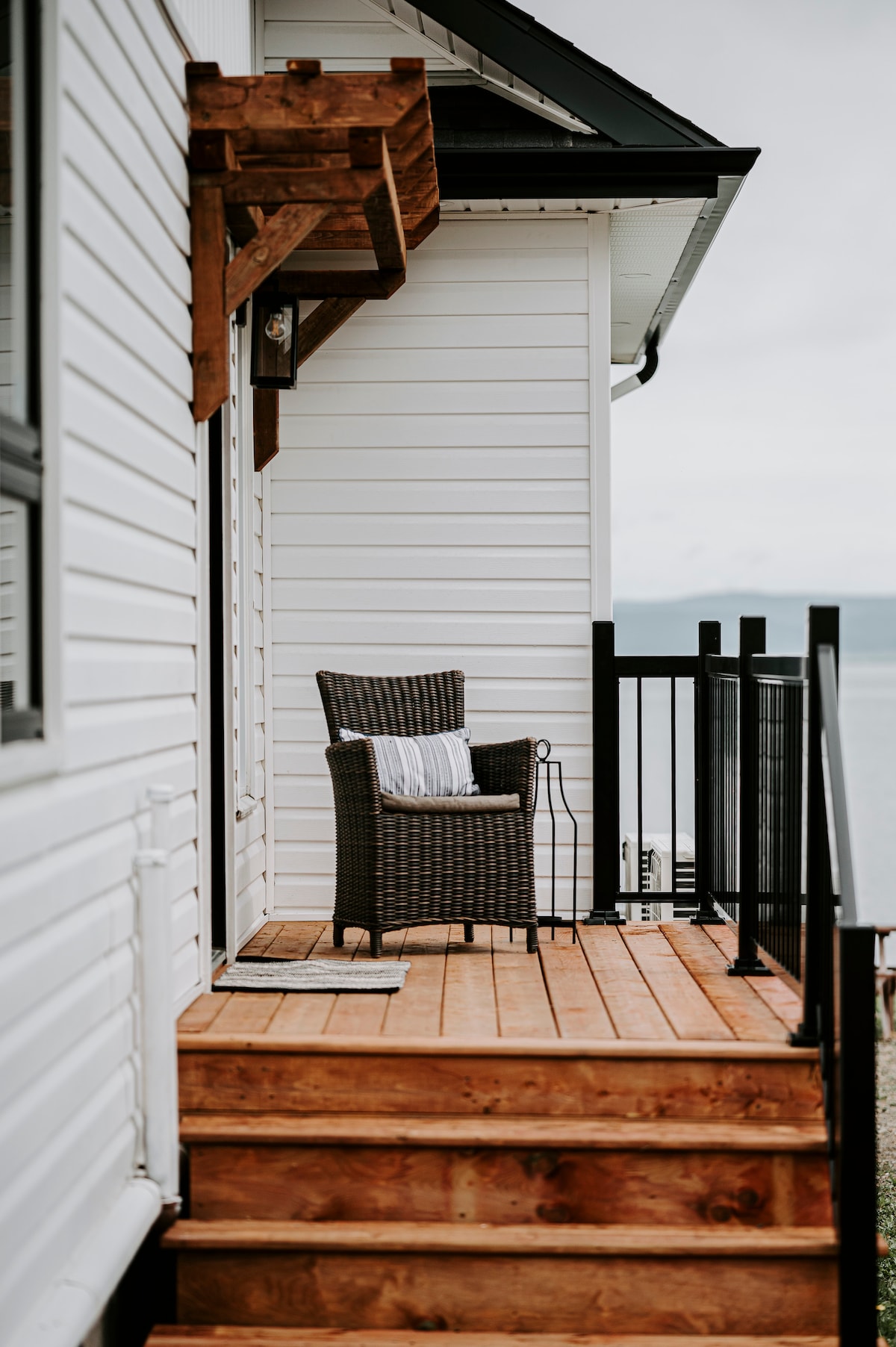 A wooden deck leads to a coastal view, featuring a single woven chair with a cushion. A side table is positioned beside the chair, offering a spot for drinks or books. The surrounding railing ensures safety and enhances the outdoor experience.