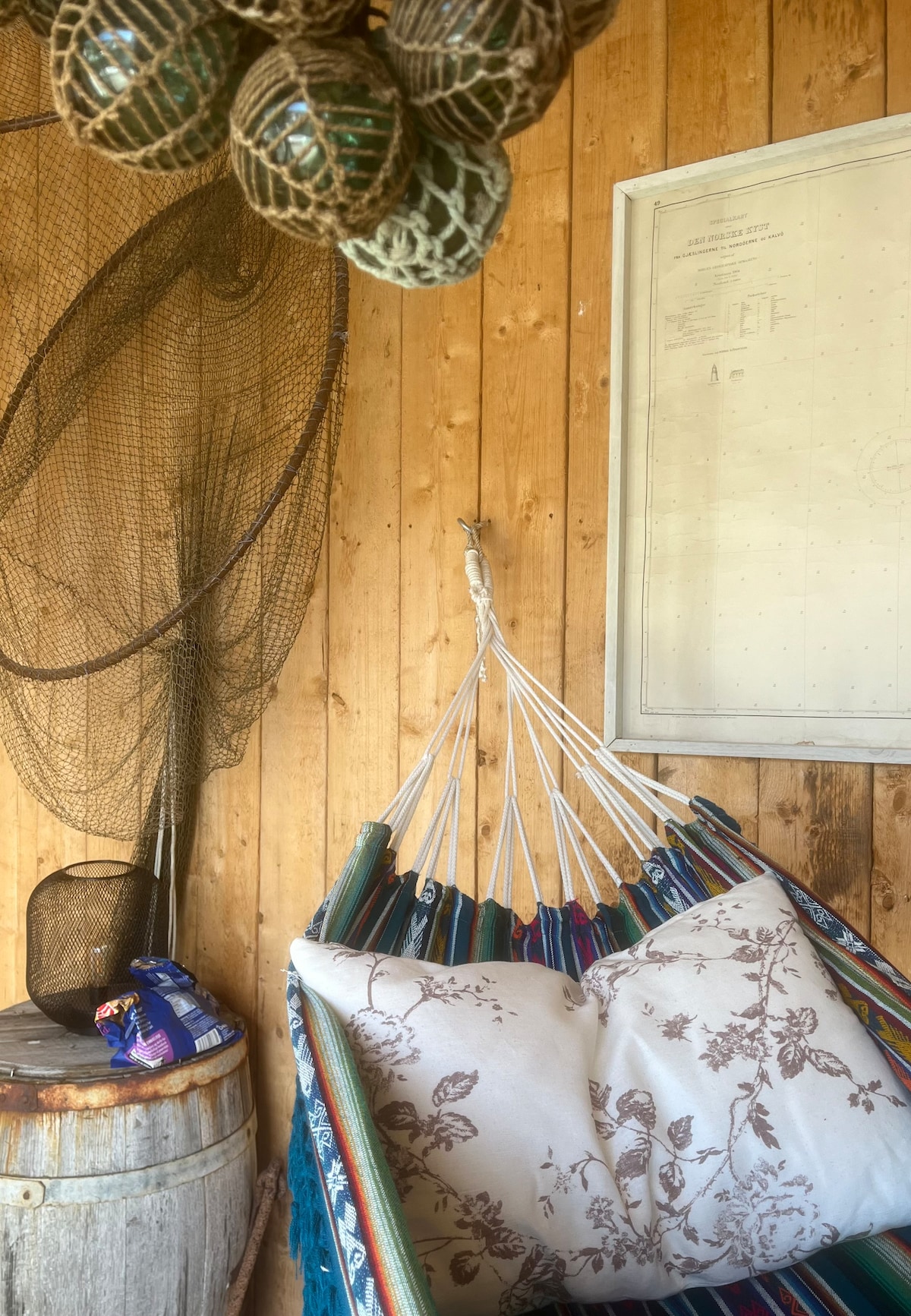 A cozy corner of the space features a colorful hammock adorned with a floral cushion. Above, decorative fishing nets and a vintage map are displayed against warm wooden paneling. A wooden barrel adds rustic charm, with a small bag resting nearby.