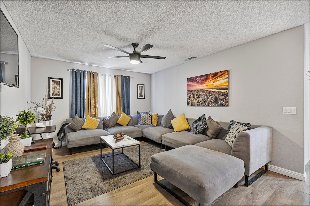 A spacious living room is presented with an oversized sectional sofa arranged around a coffee table. Bright yellow pillows add a pop of color. Natural light filters through sheer curtains, and a ceiling fan is mounted above, accompanied by framed artwork on the walls.