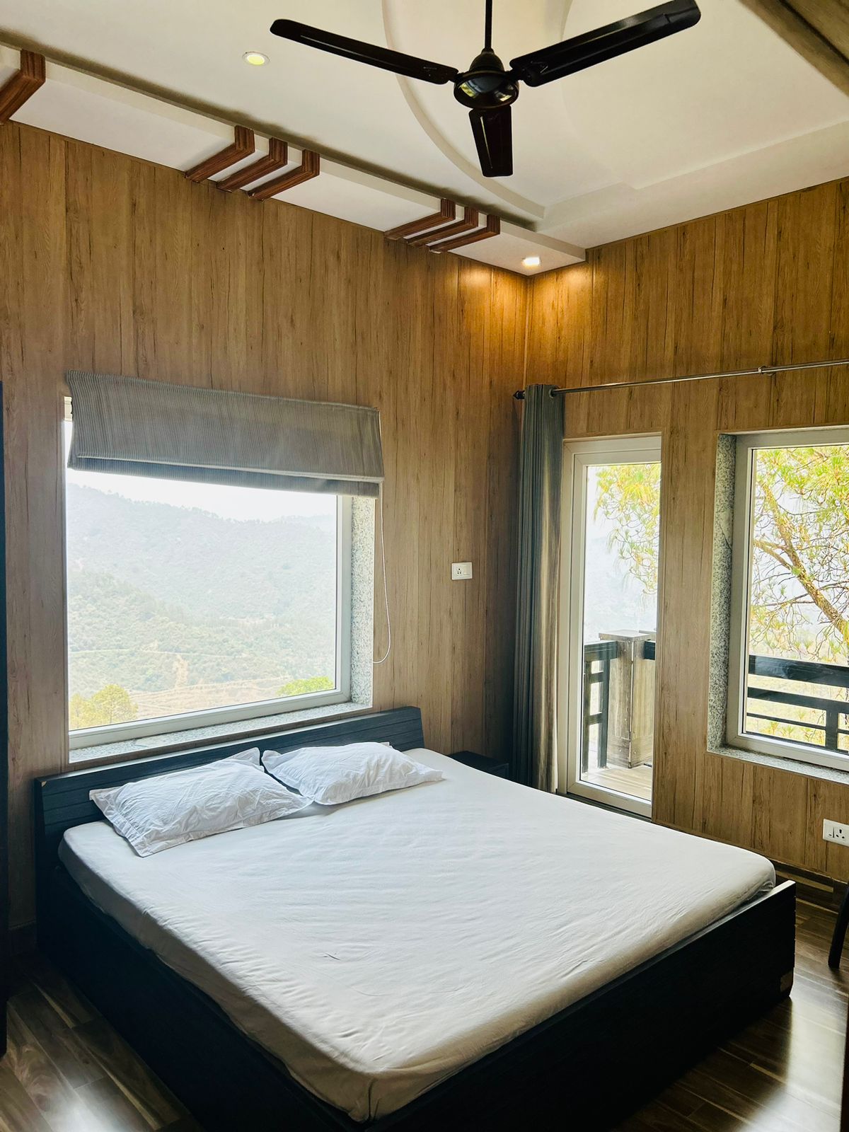 A comfortable bedroom features a spacious bed situated under a ceiling fan. Large windows provide natural light and offer views of the surrounding mountains. Wooden paneling adds warmth to the space, complemented by simple, unobtrusive decor.