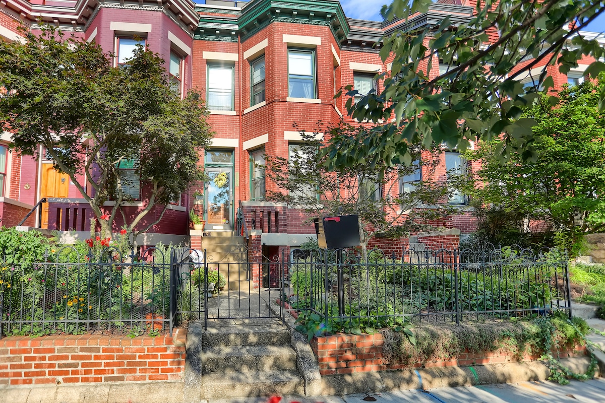 The exterior of a red brick townhouse is displayed, featuring intricate architectural details and a landscaped garden. A charming wrought-iron fence surrounds the property, with steps leading up to the front door. Vibrant green plants and blooming flowers add a fresh touch.