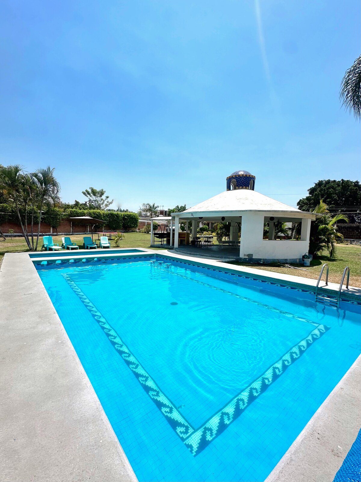 A clear blue pool is surrounded by a spacious concrete deck. Lounge chairs in soft colors are placed along the edges. A white gazebo with a thatched roof appears nearby, offering shaded seating options. Lush greenery complements the outdoor space, enhancing the overall setting.