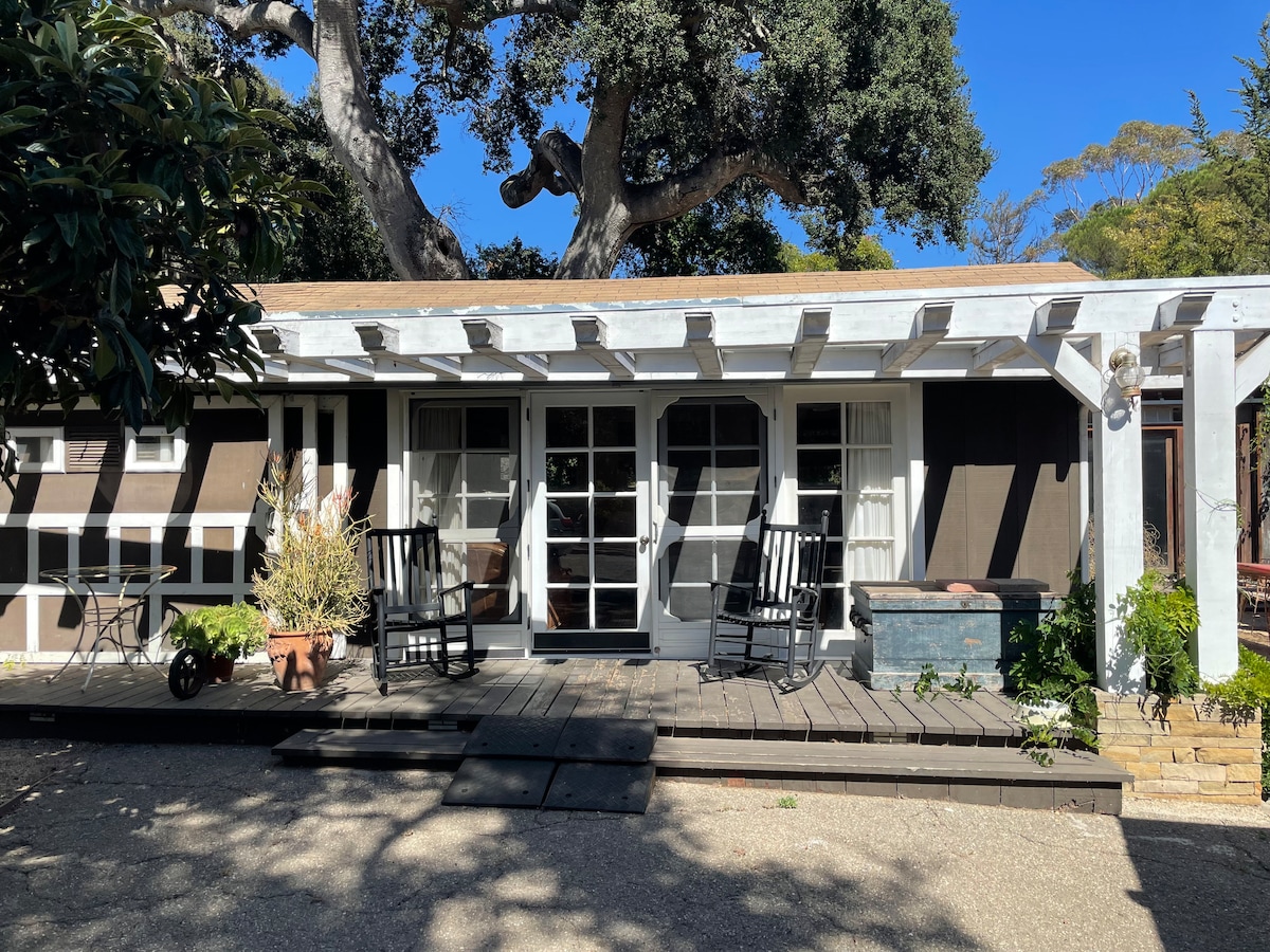 A charming cottage exterior features a welcoming porch area with two rocking chairs. Large windows allow natural light to fill the interior. The entry is framed by a pergola and is surrounded by greenery, including potted plants and a blue decorative chest.