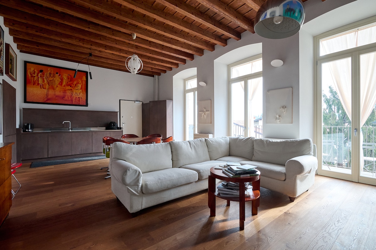 A spacious living area showcases a large white sectional sofa and a round coffee table, with natural light flooding the room through tall windows. Vintage art adorns the walls, and a modern kitchen area is visible in the background.