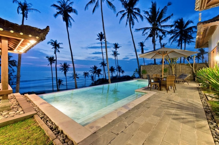 A private infinity swimming pool overlooks Balian Beach, surrounded by tropical palm trees. An outdoor dining area with chairs and a table is positioned beside the pool, providing a relaxing space for enjoying views of the sunset and ocean.