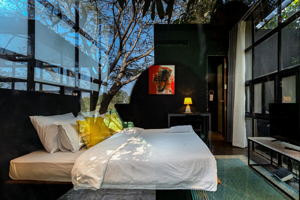 A spacious bedroom is depicted with large windows allowing natural light to fill the space. The bed is dressed in white linens with accent pillows. A small work desk and chair are visible, along with a television, enhancing the room's functionality.