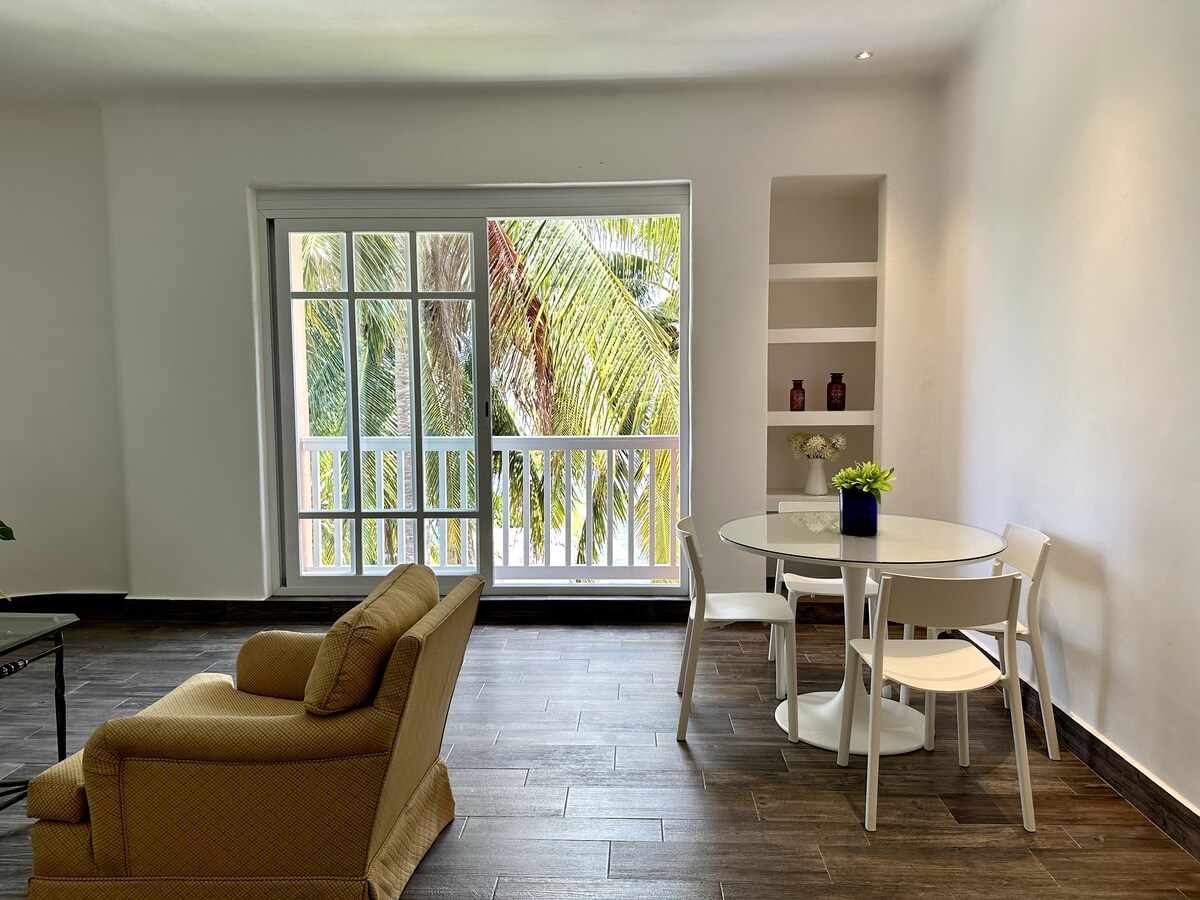 A bright and airy living space features a cozy armchair and a round dining table surrounded by white chairs. Large windows allow natural light to flood the room, offering views of lush greenery outside.
