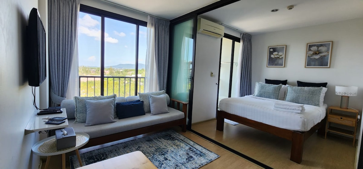 A modern living area features a spacious sofa and a large bed, complete with light-colored bedding. Floor-to-ceiling windows provide views of the surrounding landscape, while soft curtains frame the scene. Two art pieces adorn the walls, adding subtle elegance.