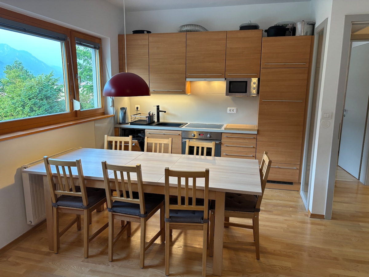 A functional kitchen space is presented, featuring light wood cabinetry and a countertop with an integrated stove and microwave. A round pendant light provides illumination over a sizable dining table surrounded by six wooden chairs. Large windows offer views of the nearby mountains.