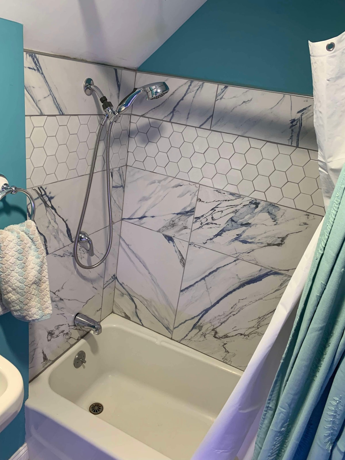 The bathroom features a compact tub with a shower above, surrounded by elegant marble and hexagonal tile work. A light blue accent wall complements the space, while a textured towel hangs conveniently on the wall.