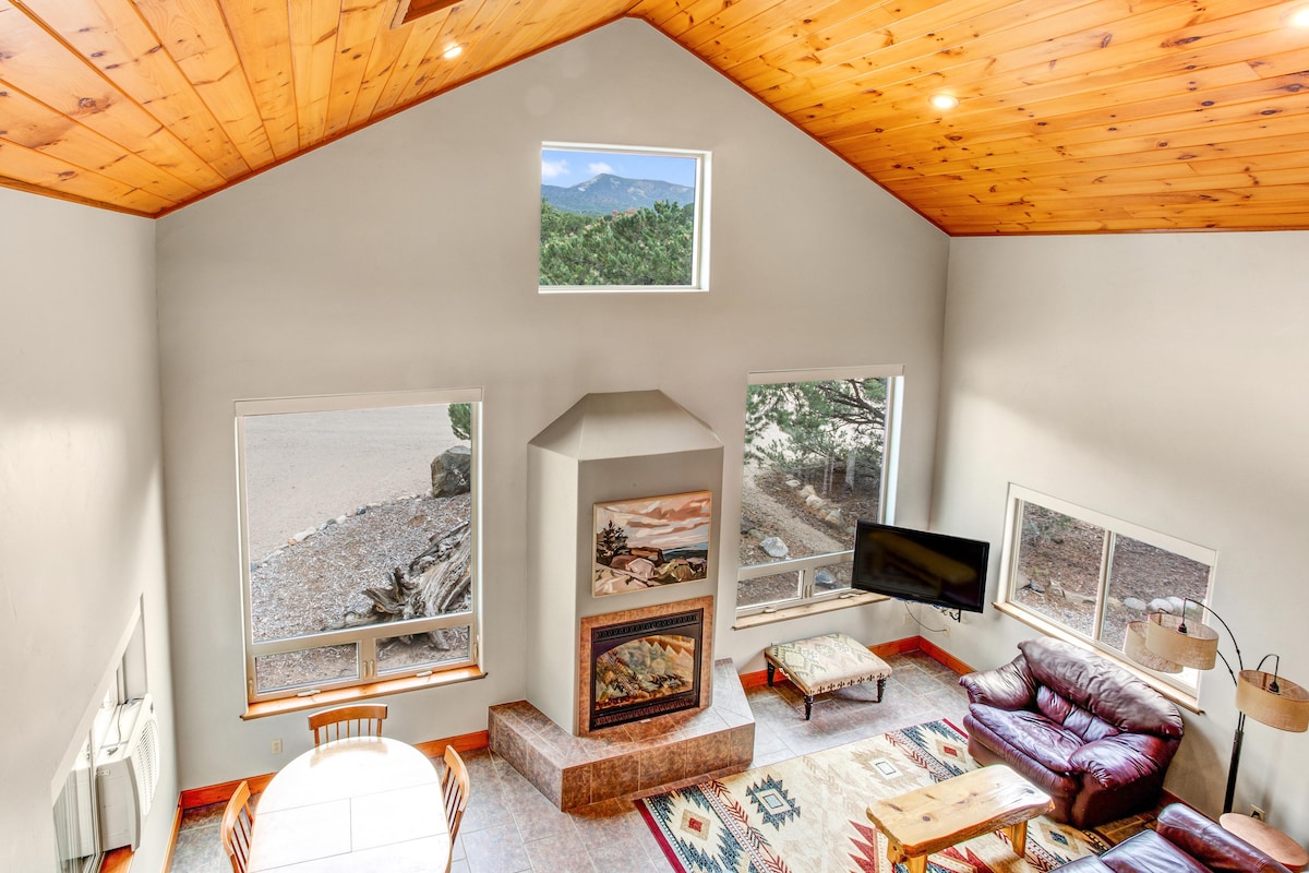 A spacious living area features a cozy fireplace at its center, surrounded by large windows allowing natural light and showcasing views of the outdoors. A leather sofa and a wooden dining table are visible, and a mounted television adds to the inviting atmosphere.