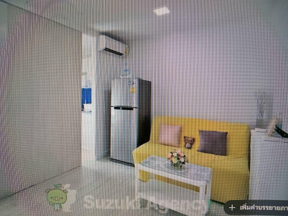 A bright living space features a compact yellow sofa paired with two pillows and a small floral arrangement on a white coffee table. A refrigerator is visible in the background, positioned next to a light-colored wall.