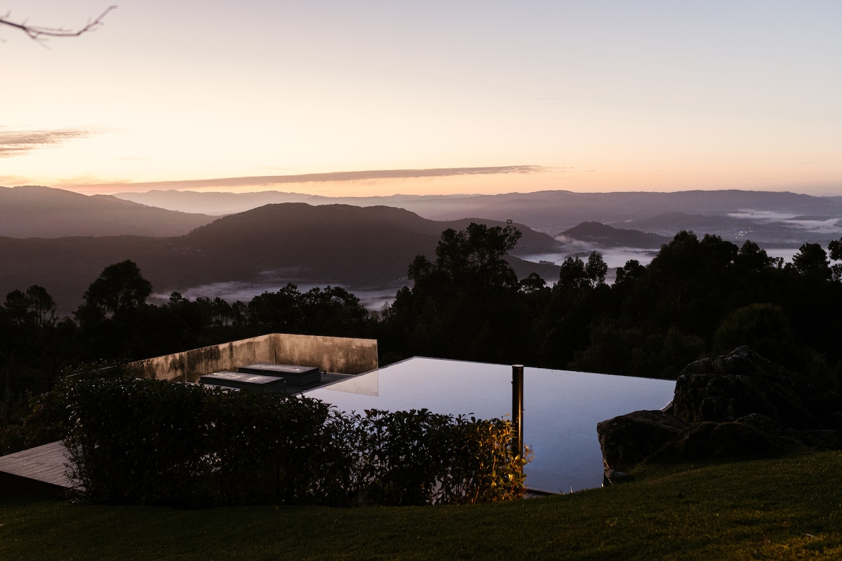 An infinity pool overlooks a vast landscape, where rolling hills meet a soft morning sky. The area is surrounded by lush greenery, providing a sense of tranquility and connection to nature.