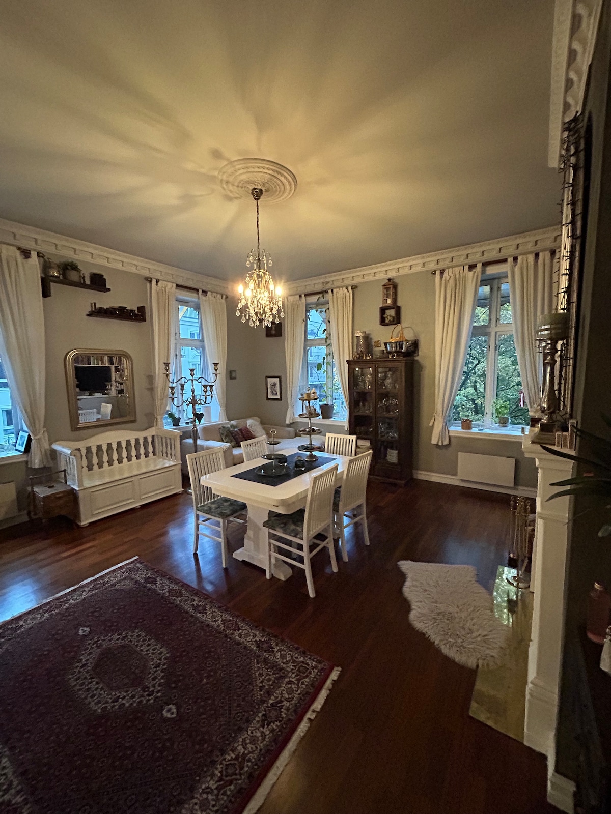 A dining area is presented with a round table surrounded by four white chairs. Large windows with sheer curtains allow natural light to fill the space, showcasing a chandelier overhead. A display cabinet and an ornate mirror enhance the room's elegant atmosphere.
