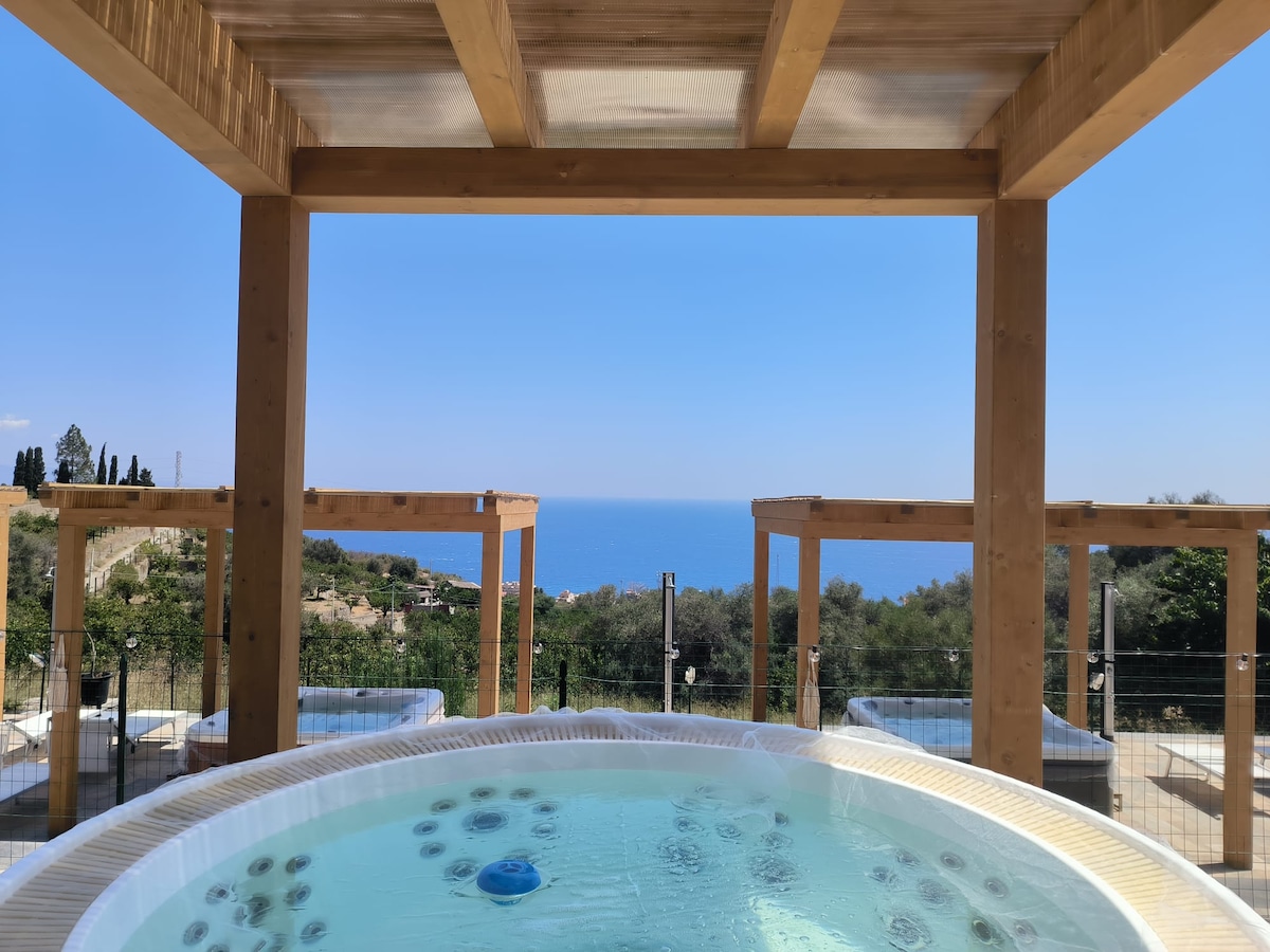A private jacuzzi is enclosed within a wooden pergola, offering panoramic views of the sea and surrounding landscape. The clear water of the jacuzzi reflects the blue sky, creating a peaceful outdoor retreat. The area is surrounded by a safety railing, enhancing the serene atmosphere.