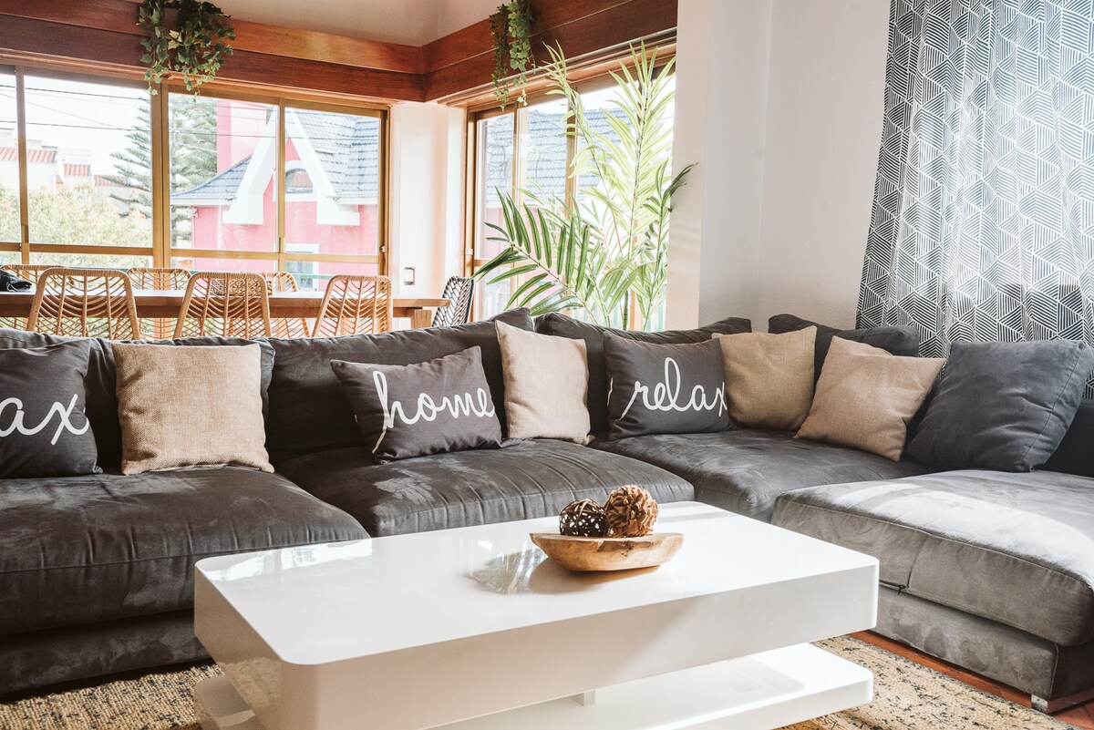 A spacious living area features a large sectional sofa adorned with decorative pillows displaying the words 'relax' and 'home.' A white coffee table with a natural wood bowl filled with pinecones is positioned centrally, complemented by a backdrop of large windows revealing the outdoor greenery.